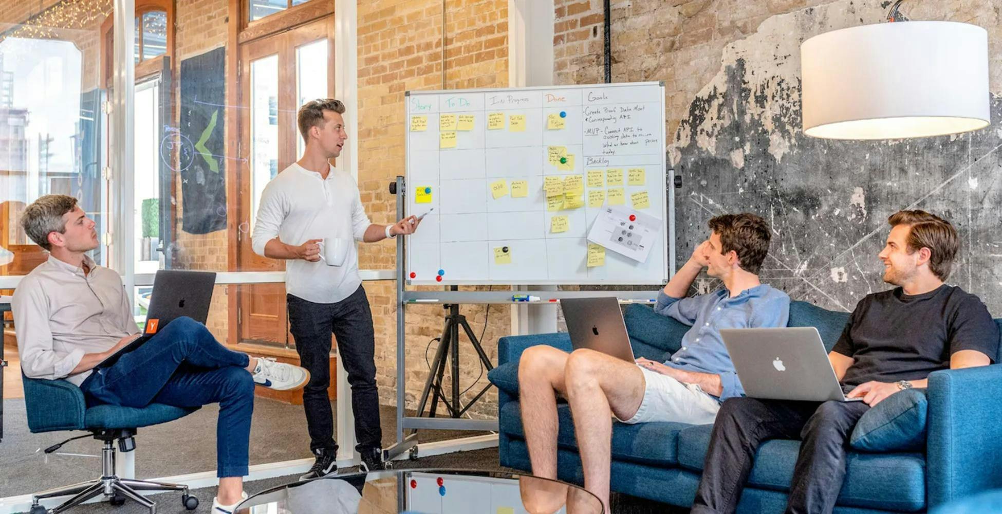 3 men sitting around looking at another man presenting on a whiteboard
